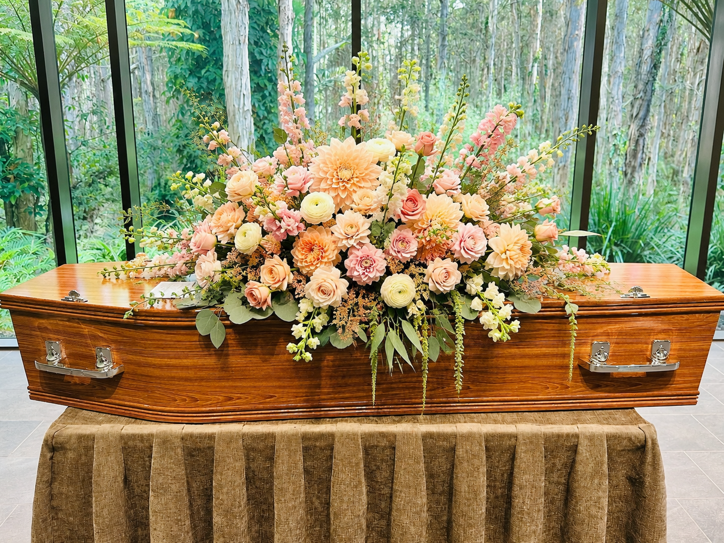 A large, elaborate floral arrangement in soft peach, pink, cream, and green tones, displayed across the top of a polished wooden casket resting on a draped table. 